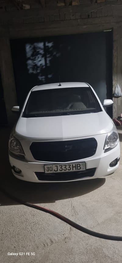 A white Chevrolet Cobalt is parked indoors, facing forward, in what appears to be a garage or workshop. The car's license plate, with the characters "30 J 333 HB" and a small Uzbek flag, is clearly visible on the front bumper. The interior of the car shows a beige-colored dashboard and seats. The environment is dimly lit, with shadows of tree branches cast on the dark background wall behind the car. The floor is concrete, and a black and red cable runs across it in the foreground. The overall scene suggests the vehicle is located in Ingichka, Uzbekistan, based on the license plate and typical automotive setting.