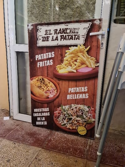 A vertical, rectangular advertising sign for "EL RANCHO DE LA PATATA" is positioned on a reddish-brown tiled ground, leaning against a white door frame. The sign features a weathered wood-effect background with three food items depicted: a pile of french fries ("PATATAS FRITAS") with yellow and pink sauces on a wooden plate, a dish containing what appears to be a halved, stuffed potato with various toppings ("PATATAS RELLENAS"), and a salad ("NUESTRAS ENSALADAS DE LA HUERTA") with a "GLUTEN FREE SALSAS" badge. To the right of the sign, part of a metallic, light-colored object, possibly a folding chair or a stand, is visible. Behind the sign, a white door frame with a glass panel is present, with a white door handle and lock mechanism visible on the right side of the door. The immediate surroundings suggest an outdoor or semi-outdoor commercial establishment in the city of El Campello, Spain.