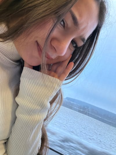 A close-up, angled photograph shows a young female with long brown hair and fair skin, looking directly at the viewer with a faint smile. Her right hand, with fingers curled, rests against her cheek. She is wearing a light-colored, ribbed turtleneck sweater. The background, visible through a window, depicts a body of water with a subtly rippled surface, backed by distant, undulating land or mountain formations under an overcast or light blue sky. This scene is located in Obermodern-Zutzendorf, France.