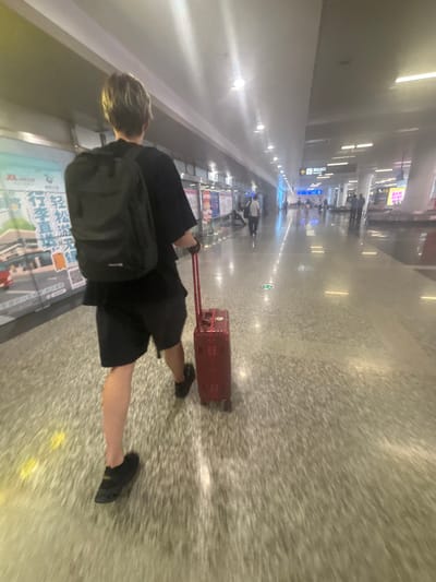 A person with blond hair, wearing a black t-shirt, black shorts, and black shoes, walks away from the camera, pulling a red suitcase with a black handle. The person also carries a black backpack. The scene appears to be an indoor walkway, likely an airport or train station, with a polished, speckled floor. Fluorescent lights illuminate the ceiling. In the background, there are signs, other people walking, and some displays on the walls. One sign with Chinese characters and an arrow points to the left, labeled "Arrival Exit". The location is identified as Xinwu District, China.