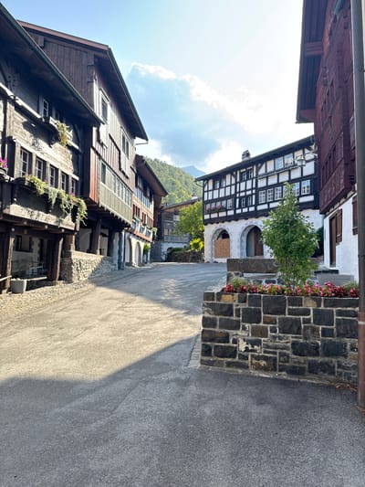 The image depicts a street scene in the historic village of Grabs, Switzerland. The street is paved with asphalt and stone, with a prominent shadow cast across it from the right side. On the left, a traditional wooden building with multiple stories and balconies adorned with flowers dominates the foreground. To the right, another building with a white facade and dark timber framing, characteristic of Swiss architecture, is visible. This building features arched entrances on the ground floor. In the center of the scene, a low stone wall enclosing a planter box with pink flowers and a small tree is positioned. Further into the background, more traditional buildings and lush green hills under a bright, partially clouded sky can be seen. The overall impression is one of a peaceful, well-preserved European village setting.