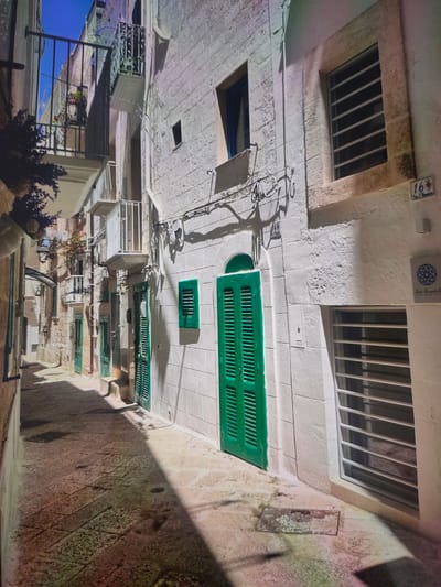 The image depicts a narrow cobblestone alleyway between two buildings with whitewashed walls. The buildings feature several windows and doors with green shutters, typical of Mediterranean architecture. Balconies with ornate metal railings extend from the upper floors. The alley is partially covered in shadow, with sunlight illuminating the cobblestones and parts of the buildings. There is a small sign with the number "16" on the right building, and a decorative circular emblem below it. The overall impression is of a historic and charming European street scene. The image is not located in Zrenjanin, Serbia, as the architectural style is inconsistent with the region.