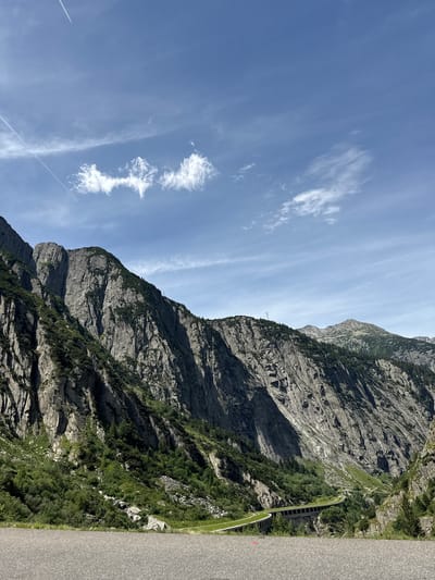 The image shows a scenic view of a mountain landscape in the vicinity of Andermatt, Switzerland. In the foreground, a paved road surface is visible. Beyond the road, a steep, rocky mountain face dominates the left side of the frame, covered in patches of green vegetation. Further into the scene, a winding road with a concrete barrier and overhead structure curves through a valley. The distant background features more mountain peaks and a bright blue sky with wispy white clouds and contrails from an airplane. The overall impression is one of a natural, mountainous environment with man-made infrastructure integrated into the landscape.