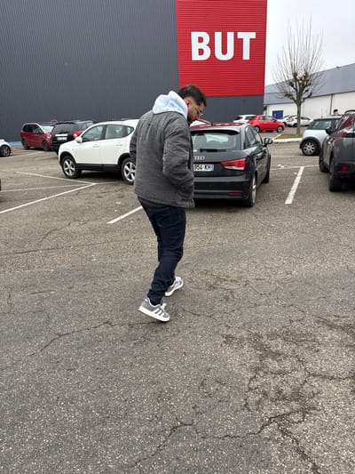 The image depicts an outdoor asphalt parking lot in front of a BUT furniture and home appliance store in Vendenheim, France, under an overcast sky. A male individual is seen from behind and slightly to his left, walking across the cracked asphalt surface, heading away from the camera. He wears a grey long-sleeved jacket over a light blue and white patterned hoodie, dark blue trousers, and grey and white Adidas sneakers. His hands are in his pockets, and his head is slightly downcast.

Behind the man, a black Audi A1 is parked with its rear facing the viewer, displaying a French license plate "954-KH". To the left of the Audi, a white Nissan Qashqai SUV is parked. Further left and behind the Nissan, a red multi-purpose vehicle is visible. The background features the large, dark grey corrugated metal facade of the commercial building, with a prominent red panel centered at the top, bearing the large white capital letters "BUT". A bare deciduous tree stands in the mid-ground to the right of the parked Audi. White painted lines delineate parking spaces on the grey, weathered asphalt.
