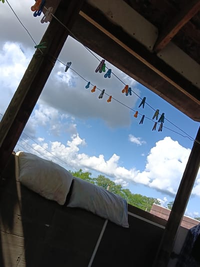 The image depicts a view from a wooden structure, likely a veranda or an outdoor shelter, looking up at a bright blue sky with scattered white clouds. Several colorful clothespins are attached to a clothesline that stretches across the frame. The clothespins are in various shades of orange, blue, white, and green. Below the clothesline, two white pillows are visible, resting on a dark surface. In the background, green trees and the rooftops of buildings can be seen. The overall scene suggests a casual, domestic setting, possibly during daytime, with laundry hanging out to dry. The image is shot with a slight tilt, giving a dynamic perspective of the sky and the surrounding elements. This scene is consistent with a typical residential area in Vityazevo, Russia, a resort town on the Black Sea coast, known for its sunny climate and outdoor activities.