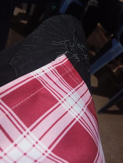 The image is a close-up shot of a person's legs, wearing dark-colored pants with some decorative stitching. A piece of fabric with a red and pink plaid pattern is draped over the pants, partially obscuring them. The plaid fabric appears to be soft and possibly part of a garment or accessory. The background is dark and out of focus, with hints of blue and brown, suggesting an indoor setting with furniture, possibly chairs. The lighting is dim, creating shadows and a somewhat moody atmosphere. There is no visible text in the image. The scene is static, with no clear activity or event taking place, focusing solely on the textures and patterns of the clothing. The location is specified as Ukpor, Nigeria, though there are no specific visual cues in the image to confirm this.