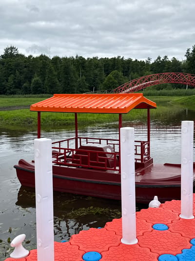 A red boat with an orange canopy is docked on a body of water. The boat has a railing and seats. In the foreground, there is a floating dock made of red and blue interlocking pieces with white posts. Behind the boat and dock, there is a line of green trees. Further in the background, a red arched bridge spans across the water. The sky is overcast. The location is in Bruzdeilynas, Lithuania.