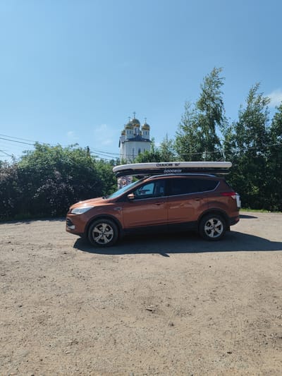 A stationary, orange Ford Kuga SUV is parked on a gravel surface in front of a lush green hedge and trees. The vehicle has two stand-up paddleboards secured to its roof rack. In the background, a white church with golden domes is visible, along with a clear blue sky. The scene appears to be an outdoor setting during daylight.