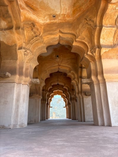 A ground-level, interior shot depicts a deep, diminishing corridor, angled slightly upward toward a distant opening revealing an exterior stone structure. The corridor's left side features an elaborate, ornate wall with repeating, scalloped archways in a warm, earthy tone. These archways are stacked in layers, creating a sense of depth and intricacy. The wall transitions to a cooler, pale tone further into the corridor. The right side of the corridor has a smooth, lighter-colored wall with horizontal lines suggesting steps or tiers leading upward. Overhead, a series of simple, rectangular beams span the corridor, providing a structural framework. The lighting is uneven, with the foreground walls being more intensely lit than the darker, recessed areas. In the distance, the opening to the exterior reveals a section of stone construction. The image is from Hampi, Kamalapura, India.