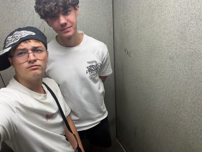 Two young men are posing for a photo in what appears to be a confined space with textured grey walls, possibly an elevator or a small room. Both individuals are wearing white t-shirts. The man on the left has curly brown hair and is looking towards the camera with a neutral expression. He has some acne on his face. The man on the right is wearing glasses with thin, round frames and a black baseball cap with a flame design. He is also looking at the camera. A dark strap is visible across the shoulders of the man on the right, suggesting it might be a bag strap. The lighting in the scene is somewhat dim and casts shadows, giving it a casual, unposed feel. There are some faint markings on the wall to the left, including what looks like handwritten text. The overall mood is relaxed and informal. The location context is Dorsten, Germany.