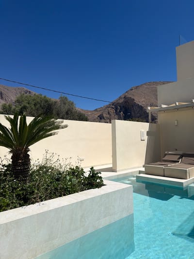 This is a medium-shot, eye-level, outdoor, daytime photograph of a private swimming pool area adjacent to a building, set against a backdrop of arid, rocky mountains. The scene is located in the Municipal Unit of Thira, Greece.

The foreground features a pristine, bright blue swimming pool, with clear water and visible tiling on the bottom. To the left, a portion of the pool's edge is visible, constructed from light-colored, textured stone tiles. A lush green plant, possibly a cycad, with dark, spiky fronds, is planted next to the pool. Behind the plant is a low wall, also tiled in the same light stone. Beyond this, a tall, pale yellow wall encloses the area.

In the mid-ground, a modern building with clean, white architectural lines is situated to the right. It has an overhanging roof structure and features a recessed section with two flat, brown lounge chairs arranged side-by-side. A smaller, off-white wall is positioned in front of this building, also part of the outdoor space. A small, white rectangular object, possibly a light fixture or intercom, is mounted on this wall. Further back, a tall, pale yellow wall continues, and on its surface, a framed document or sign is visible.

The background is dominated by rugged, brown, rocky mountains, with sparse vegetation clinging to their slopes. The sky above is a vibrant, cloudless blue. Overhead, power lines are strung across the sky, supported by unseen poles. The overall impression is one of a luxurious, tranquil vacation setting in a Mediterranean environment. There are no people or animals present in the image.