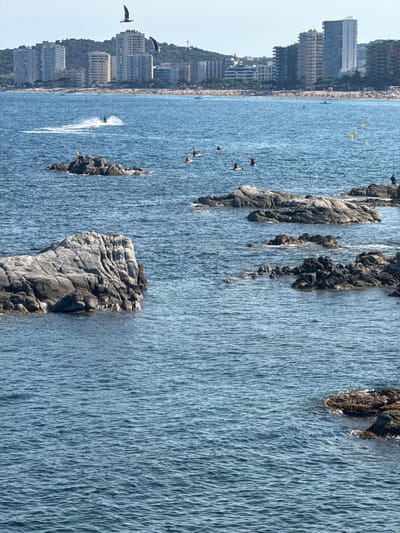 The image depicts a coastal scene with the city of Platja d'Aro in the background. The water is a deep blue with small waves. Several rocky outcroppings are visible in the foreground and middle ground, with some people in kayaks and a jet skier in the distance. The city, situated on a hillside, features a cluster of multi-story buildings, including residential apartments and hotels, overlooking a beach with scattered sunbathers. Two seagulls are captured in flight in the upper portion of the image. The overall impression is of a sunny day at a popular Mediterranean resort town.