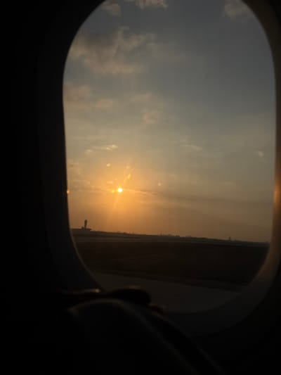 The image depicts an exterior view captured from within an aircraft cabin through an oval-shaped window, positioned at Ben Gurion Airport (TLV) near Yehud Monosson, Israel, during either a sunset or sunrise. The sky dominates the upper portion of the frame, showcasing a bright orange-yellow sun with radiating lens flares in the upper-left quadrant. The sky color transitions from intense orange-yellow near the horizon to a paler blue-grey further up, with scattered white cloud formations present. Below the horizon, a flat airfield surface is visible, consisting of dark ground material and a lighter, horizontal strip, indicative of a runway or taxiway. A tall, slender air traffic control tower, silhouetted against the bright sky, stands prominently in the mid-ground towards the left. A faint, distant urban skyline is discernible along the far horizon to the right. The dark, curved edges of the aircraft's window frame define the perimeter of the view, and an indistinct, dark object, possibly part of the aircraft's interior or a passenger's personal item, occupies the bottom foreground. No individuals, specific actions, or direct interactions are visible in the scene.