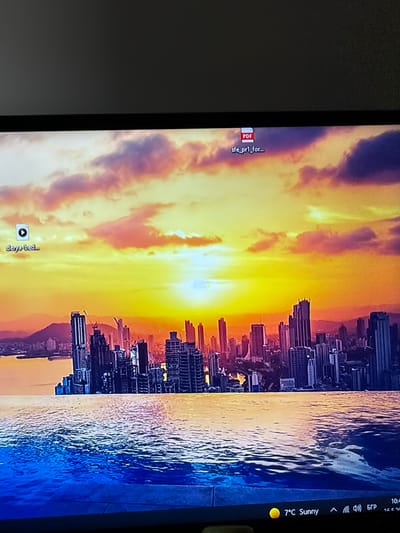 The image shows a computer screen displaying a desktop with a sunset/sunrise cityscape wallpaper reflecting in water. There are two desktop icons: a video file named "derya-bed..." and a PDF document named "sfe_pr1_for...". The taskbar on the right-hand side indicates the temperature is 7°C, the weather is sunny, and shows the time as 10:4_. There are also icons for speaker volume and language. The location is not depicted in the image.