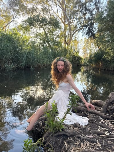 A woman with long, curly brown hair and a crown of green leaves sits on the exposed roots of a tree at the edge of a body of water. She wears a white, strapless dress that flows around her. Her left leg is submerged in the water, with her bare feet visible. The water reflects the sunlight and the surrounding trees. The background consists of dense green vegetation and tall trees, suggesting a natural, possibly wooded, setting. The lighting indicates it is daytime, likely late afternoon, given the warm tones of the sun. The location is described as being in the city of Katzrin, Israel.