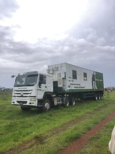 A mobile training platform truck, identified as "EDUCATIONAL" and bearing the inscription "FEDERAL GOVERNMENT OF NIGERIA", is parked in a grassy field. The truck's cargo container is painted white and green and features several windows. Text on the container indicates it is a "MOBILE AUDIO-VISUAL TRAINING PLATFORM". Two outdoor air conditioning units are mounted on the exterior of the container. The scene appears to be set in Farin Yashe, Nigeria, with some distant buildings visible. The sky is overcast with clouds. Two individuals are faintly visible walking in the background near the buildings. The ground is covered in green grass and a dirt path.