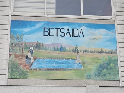 The image is a photograph of a painting titled "Betsaida" displayed on a building in Osorno, Chile. The painting depicts a landscape with a rectangular body of water in the center. To the left of the water, a person sits in a wheelchair next to a standing figure dressed in a nun's habit. On the right side of the water, a person in a white robe stands and appears to be gesturing towards the water, with rays of light emanating from the sky above them. The word "BETSAIDA" is painted in large black letters across the top of the artwork. The painting is affixed to a building with white horizontal siding and a window above it.