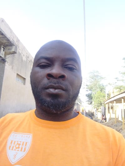 A bald, middle-aged Black man poses close to the camera. He has a dark beard with some gray hairs and wears an orange t-shirt featuring a logo of a soccer ball and "SPORTING". He stands outdoors; behind him are simple concrete buildings with peeling paint, trees, and a street scene with people visible in the distance. The sky is bright and clear.