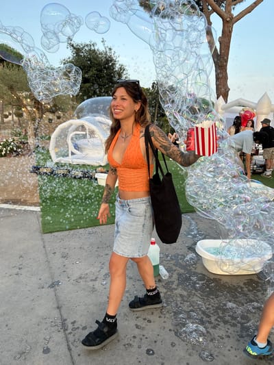 In Sant Vicenç De Montalt, Spain, a woman is captured mid-stride, holding a red and white striped bucket overflowing with popcorn. She wears an orange halter top, a light blue denim skirt, black sandals with socks, and sunglasses perched on her head. Her arm is adorned with tattoos, and she carries a black tote bag. She is walking on a concrete path, with a large display of bubbles surrounding her. To her left, a patch of artificial grass features a white tent-like structure. In the background, several people are visible, some standing near a table, and a few balloons are scattered around. The scene appears to be an outdoor event, possibly a festival or party, with a festive atmosphere suggested by the bubbles and popcorn.
