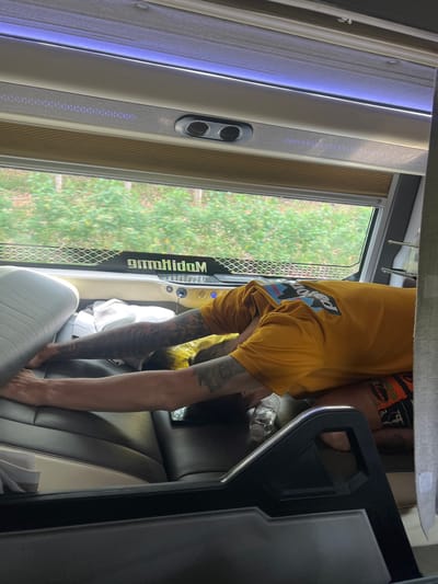 An interior view shows a person bending over in what appears to be a passenger seat of a bus or large van. The person wears a yellow t-shirt with a "Patagonia" logo and has tattoos on their arms. The seats are upholstered in a light gray and dark gray material with white accents. A black panel with a metallic trim is visible to the right of the seat, and a window on the left shows green trees and vegetation outside. Part of the word "Kiah" is seen reversed on the window frame. A water bottle is visible near the person. The setting implies a vehicle traveling through Kon Ra Kiah, Vietnam.