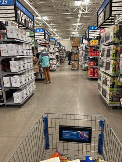 The image, oriented vertically, presents a view down a wide aisle inside a large retail store, possibly a big-box retailer. On the right side, a shopping cart is partially visible in the foreground, with a rectangular sign in its basket reading "Attention shoppers" along with a prohibited symbol.

Tall metal shelves line both sides of the aisle, densely packed with various products in boxes. On the left side, a price tag shows "$19.98" (or "$19.85") for "Everyday Low Price," with items like "Moth balls," "Lint rollers," "Organizers," and "Laundry totes" listed below. Aisle signs "H 27" and "H 25" are visible further down this side. The shelves hold numerous white-boxed goods, with some labels suggesting small appliances like coffee makers. The right side features shelves stocked with similar items, including "Hamilton Beach" microwaves, and an aisle sign "H2."

Two individuals are seen working in the aisle. Closer to the foreground on the left, a person in a light blue/green vest and brown pants is bent over, appearing to stock or organize products on the lower shelves. Further down the aisle, another person in dark clothing is near rolling carts laden with cardboard boxes, also engaged in work.

The floor is grey concrete, and the ceiling features exposed metal beams and grid structures. The scene suggests an active retail environment, likely during operational hours.