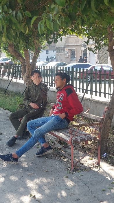 Two young males are seated on a weathered, reddish-brown metal and wooden bench under the partial shade of trees in Al-Qaboun Municipality, Syria. The male on the left wears a dark olive green zipped tracksuit and dark sneakers; his right hand is raised, covering his face from the nose upwards, and his left hand rests on his lap. The male on the right wears a red long-sleeved hoodie with black shoulder sections and a white graphic on the chest, light blue jeans, and dark blue sneakers; his right hand is in his jeans pocket, and his left arm rests on the back of the bench as he looks to his right.

The ground in the immediate foreground is a concrete surface, partially illuminated by sunlight and dappled with shadows from overhead tree foliage, with scattered dry leaves and a discarded blue soda can near the bench's right leg. Behind the bench, a dark metal fence with vertical bars separates the seating area from an asphalt street. Several vehicles are parked along the street, including a silver sedan and a red hatchback. Across the street, old, light-colored multi-story buildings with various windows and architectural details are visible, with indistinct figures present in the distance near the buildings and vehicles. The scene is illuminated by bright daylight.