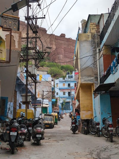The image depicts a narrow street in Jodhpur, India, with a prominent fort visible in the background. The street is lined with multi-story buildings exhibiting a mix of architectural styles and colors, predominantly featuring blues, yellows, and faded plaster. Numerous utility poles with a dense network of electrical wires crisscross the scene, indicating a common infrastructure in the area.
On the street level, a variety of vehicles are parked and in motion. Several motorcycles and scooters are the most numerous, with license plates visible on some, such as "RJ19E 6631" and "RJ19B K9758." A yellow auto-rickshaw and a light-colored truck are also present. A few people are visible walking and standing in the street, some engaged in conversations or going about their daily activities. Children can be seen playing in the distance. The ground is paved and shows signs of wear, with some puddles or wet patches. The overall atmosphere suggests a vibrant urban environment with dense living and commercial activity. The fort in the background, likely Mehrangarh Fort due to its characteristic red sandstone and commanding presence, is a defining landmark of Jodhpur. The sky is overcast, suggesting a cloudy day.