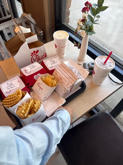 Here's a concise description of the image:
A high-angle shot depicts a meal from Chick-fil-A, including waffle fries, sandwiches, and drinks, arranged on a black serving tray on a light brown woven table. The presence of the name "LAURA 4921 DINE IN" on several items suggests the location is the Chick-fil-A at 4921 West Southlake Boulevard, Southlake, Texas, United States of America. A person wearing a light blue long-sleeved shirt is partially visible in the lower right corner, placing their hand near the tray. A small vase with artificial flowers sits next to the window in the background, near the camera and the drink. A digital camera also sits on the table next to the window. A black chair is visible in the background, suggesting this is an indoor restaurant setting.