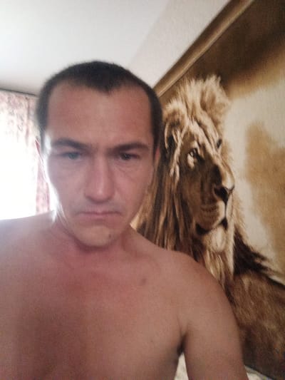 A man is in the foreground, shirtless, with dark hair and fair skin. He is looking at the camera with a neutral expression. In the background, there is a tapestry or blanket depicting a lion. The scene appears to be indoors, possibly in a bedroom, with a curtain visible on the left side. The lighting is somewhat dim, with light coming from the left. The location is identified as Кадзінскі сельскі Савет, Belarus.
