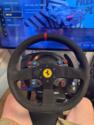 The image displays a Ferrari racing wheel in front of a television screen. The wheel features a black suede-like material on the rim, a yellow Ferrari logo, and various buttons labeled "MAIN," "RADIO SCROLL," "PIT," and "WASH." The screen shows a racing game menu with options like "MEETUPS," "FREE PLAY," "RIVALS," and "PRIVATE MULTIPLAYER," with "FREE PLAY" highlighted. The phrase "ANY CAR, ANY TRACK" is visible at the top of the screen, along with an image of a race car.