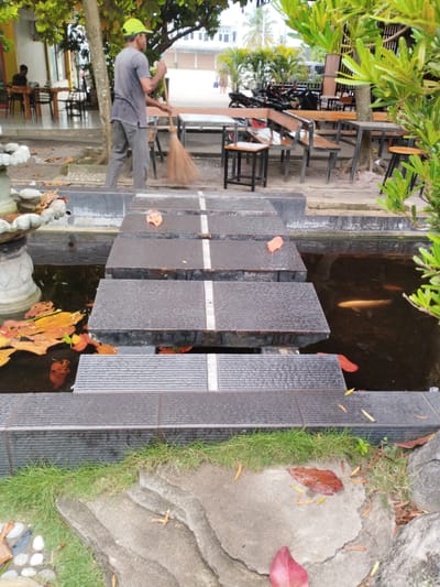 A man wearing a grey t-shirt and grey pants, and a bright green cap, is sweeping the ground with a broom. He is standing near a water feature with stepping stones that cross it. Several orange and red leaves are floating in the water. In the background, there are tables and chairs, motorcycles, and trees. The scene appears to be an outdoor seating area, possibly a cafe or restaurant, in Payameuneng, Indonesia. The environment is somewhat unkempt, with fallen leaves scattered around the area. The man's action suggests he is performing maintenance or cleaning duties.