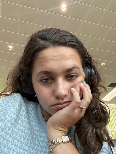 The image is a close-up, selfie-style photograph of a young woman with long, dark, wavy hair. She is wearing black headphones and a light blue patterned sweater. Her right hand is resting on her cheek, and she is wearing a gold ring and a gold watch. Her expression appears tired or bored. The background is a tiled ceiling with fluorescent lighting. The location is not specified as Azzano San Paolo, Italy, nor is there any indication of a city.
