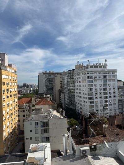 The image displays a dense urban landscape in Porto Alegre, Brazil, consistent with areas such as the Centro Histórico, Independência, or Bom Fim neighborhoods. The sky above is clear blue with scattered, wispy white clouds.

The foreground consists of various building rooftops, primarily flat, light-colored concrete or tiled surfaces, showing signs of weathering. These roofs feature cylindrical metal vents, white rectangular air conditioning condenser units, multiple white satellite dishes, and a tangle of exposed electrical cables. One prominent rooftop on the right includes a large, multi-element television antenna.

In the midground, a diverse array of buildings dominates the scene. On the left, a tall, multi-story building with a mustard-yellow facade rises, characterized by numerous small, uniformly spaced windows and external air conditioning units mounted beneath many of them. Its upper section is lighter in color. Below this, and extending towards the center, are several lower-rise residential buildings with distinct reddish-brown terracotta tiled roofs, suggesting older architecture.

To the right, a prominent, multi-story white building features numerous windows, many equipped with external white sun blinds or shutters, and multiple rows of air conditioning units. Its flat roof supports an array of communication infrastructure, including several cellular antennas and radio masts. Behind this white building, a taller structure with a dark, possibly glass or dark-paneled facade is visible. Scattered green tree canopy is discernible between the buildings, indicating street-level vegetation.

The scene depicts a static urban environment. No individuals are visible, nor are any specific actions or interactions taking place. The buildings appear to be primarily residential or mixed-use, contributing to the dense urban fabric under clear daylight conditions.
