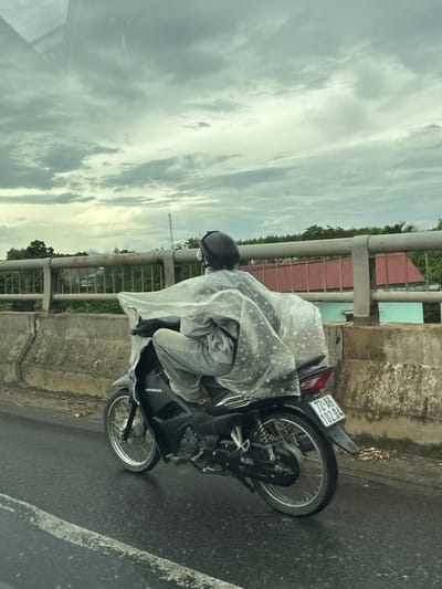 A person rides a black Honda motorcycle on a wet, paved road, likely in Nam Hải Lăng Commune, Vietnam, under a cloudy sky. The rider is wearing a gray outfit and a black helmet and is covered by a clear plastic poncho patterned with white stars. The motorcycle has a license plate reading "72-AH 102.04". A concrete barrier with a metal railing runs alongside the road, with trees and buildings visible in the background. The road surface appears to be damp, suggesting recent rain.