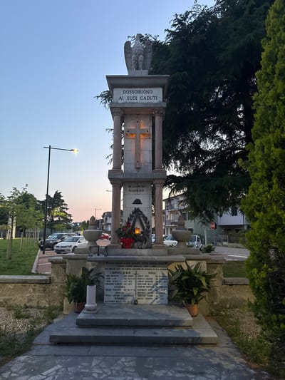 Here is a forensic description of the image:

The image depicts a war memorial in Dossobuono, a town within the city of Villafranca di Verona, Italy. The monument is dedicated to the fallen soldiers of the town, with names inscribed on the stone slab. The inscription reads "DOSSOBUONO AI SUOI CADUTI" (Dossobuono to its fallen). There are names listed under the headings of Cephalonia, Croatia, Germany, and Russia. An eagle statue sits atop one section. The surrounding environment includes trees, a street light, other graves, and parked cars. A stop sign is visible in the background.