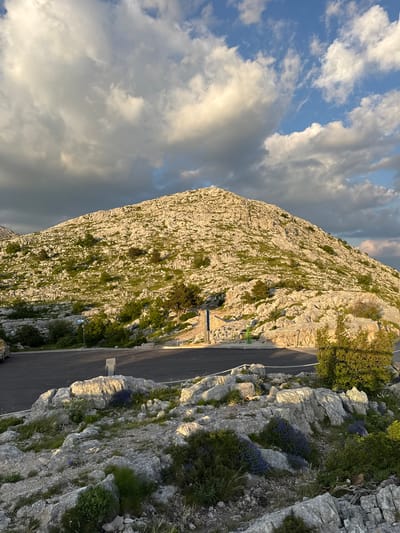 The image captures a wide-angle view of a rocky, mountainous landscape under a partly cloudy sky at what appears to be golden hour. The foreground features a paved road and parking area, with scattered rocks and low-lying green vegetation, including patches of small purple flowers. The mountain slope rises steeply, characterized by abundant gray rocks and patches of green grass and sparse trees. The lighting suggests the sun is low, casting long shadows and highlighting the textured surfaces of the mountain. In the distance, a few dark clouds are visible against the brighter sky. The scene is devoid of any people. This location is likely along the scenic route leading to or from a viewpoint within the municipality of Općina Tučepi, Croatia, known for its mountainous terrain adjacent to the Adriatic coast.
