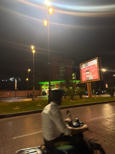 The image, taken at night, shows an urban scene in Ouahat Sidi Brahim, Morocco. A large billboard displays an advertisement in red and white tones. Adjacent to the billboard is a building with an image of a face mural painted on its side. A gas station with prominent green lighting is also visible, indicating commercial activity. In the foreground, the handlebars of a vehicle, possibly a motorcycle or scooter, are visible. Streetlights illuminate the scene, casting a yellow glow. The street has a paved shoulder and a strip of grass separating the roadway from the commercial buildings. The image is somewhat blurry, particularly in the foreground.