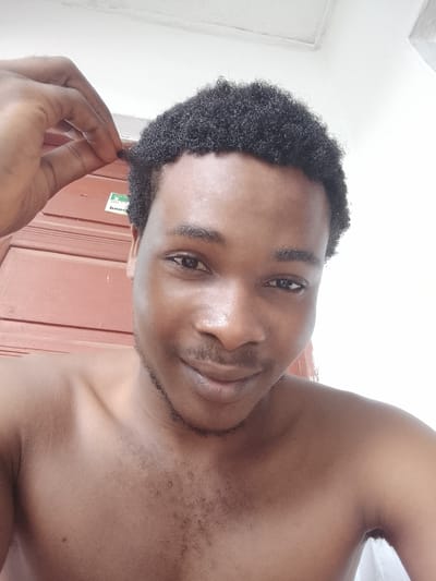 This is a close-up selfie of a young Black man. He has dark, curly hair and is looking at the camera with a slight smile. His right hand is raised, gently touching the top of his head. He is shirtless, and the background appears to be indoors with wooden paneling and a white wall. A small sticker with text is visible on the wooden paneling. The lighting is bright, suggesting daytime. The overall mood is casual and relaxed. The location is identified as Orodo, Nigeria.