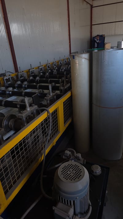 The image depicts a metalworking machine, likely a roll forming machine, in an industrial setting. Two large metal coils are positioned above a series of rollers within a yellow-framed framework. A motor is visible on the right side of the image. The background suggests the interior of a warehouse or factory with corrugated metal walls. There is a person or people near a truck in the back of the building. There is not enough information in the image to determine if the location is in Balgarevo, Bulgaria.