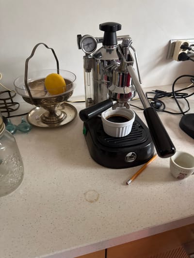 The scene depicts a kitchen countertop in the city of Le Raincy, France. The countertop is light-colored with speckled aggregate, occupying the lower portion of the frame, with light brown wooden cabinetry visible below it. A plain white wall forms the background.

On the left, a clear glass bowl with an ornate metal stand and handle, appearing tarnished, contains a single yellow lemon submerged in clear liquid. This bowl rests on a circular metal plate. Adjacent to it on the left, a textured clear glass jar is partially visible. Behind the glass bowl, two small, light blue glass objects are present.

Centrally positioned is a chrome La Pavoni lever-operated espresso machine with black handles and accents. A pressure gauge is visible on its upper left side. A small white fluted ceramic demitasse cup, filled with dark liquid identified as espresso, is positioned directly beneath the portafilter on the machine's black drip tray.

To the right of the espresso machine, a yellow wooden pencil with a pink eraser lies horizontally on the countertop. Further to the right, a small white ceramic mug with red markings is partially visible. A distinct circular brown coffee stain is present on the countertop below the espresso machine. In the upper right background, a cream-colored electrical outlet with two European sockets is mounted on the white wall, from which several black electrical cords extend.