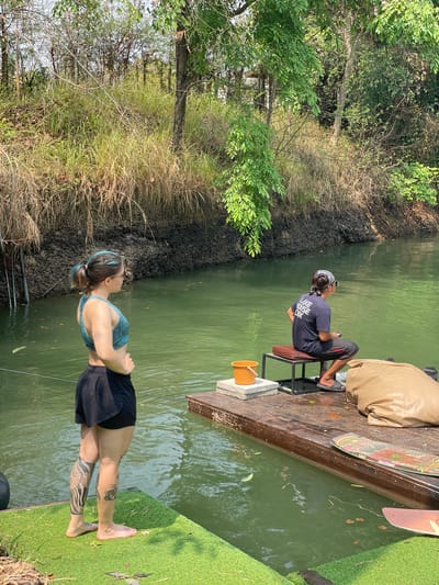The image shows a waterway adjacent to land in Tambon Nam Phrae, Thailand. A woman stands barefoot on a patch of artificial turf near the water's edge. She is wearing a sports bra and shorts. A man sits on a bench on a wooden dock, further along the waterway. He is wearing a t-shirt and pants. There is a wakeboard on the dock near him. The water is green. The banks are lined with vegetation and dark soil.