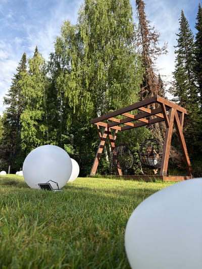 The image depicts an outdoor scene in a rural setting, likely within the Tarhankka rural district of Kazakhstan. Dominating the foreground are several large, white, spherical objects, one of which is partially visible and out of focus, suggesting they are large outdoor lights or decorative orbs. One orb in the middle ground is clearly visible, featuring a small solar panel on its surface, indicating it is solar-powered.
Behind these orbs, a wooden A-frame structure stands, constructed from dark brown lumber. Hanging from this structure are two black, wire-mesh spherical chairs, designed for seating. One of these chairs contains what appears to be a person sitting inside, though the details are indistinct.
The background is filled with lush green trees, primarily deciduous with some coniferous trees interspersed, suggesting a natural, possibly wooded environment. The sky above is a bright blue with scattered white clouds, indicating fair weather and daytime. The angle of the shot is low and tilted, looking upwards towards the trees and sky. The overall impression is one of a serene outdoor recreational or decorative space in a natural landscape.
