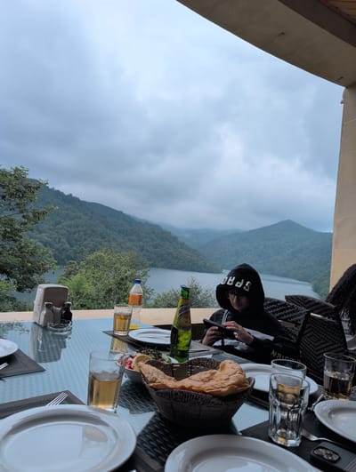 The image depicts a dining scene with a scenic view of a lake surrounded by forested mountains. In the foreground, a glass-topped table is set with plates, cutlery, glasses of amber liquid, a bottle of "NATAKHTARI" pear-flavored drink, a basket of bread, and a small bowl of salad. A person wearing a black hoodie with white lettering and glasses is seated at the table, looking at a smartphone. The background reveals a wide expanse of water, likely a lake, nestled between lush green, tree-covered mountains under a cloudy sky. The setting appears to be an outdoor restaurant or cafe terrace with a stone pillar and overhang on the right side. The location is identified as being in the city of Toğanalı, Azerbaijan.