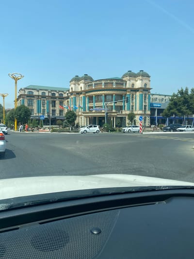 An eye-level medium shot captures a wide, paved intersection under a clear blue sky, with a prominent, ornate hotel dominating the background. The hotel, named "Hotel Orient" according to its signage, exhibits a grand architectural style with cream-colored facades, teal decorative columns, and dark green domed roofs. Flags of various nations, including those of Uzbekistan, are displayed on the hotel's exterior. Several vehicles, including a white sedan, a black SUV, and other cars, are present at the intersection. Pedestrians are visible on the sidewalks and crossing the street. The foreground is filled with the dashboard and windshield of a vehicle, suggesting the photo was taken from inside a car. The environment appears to be urban, with trees and streetlights lining the road. The scene is set in the city of Andijan, Uzbekistan.