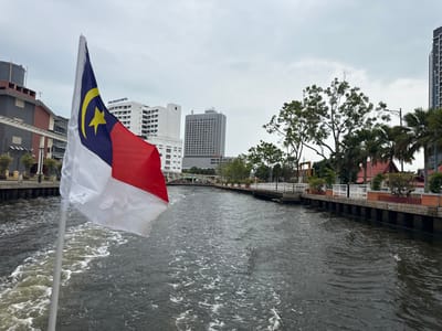 A view from a boat traveling on a canal, with a Malaysian flag prominently displayed in the foreground. Buildings line the canal, including a white building with the words "PUTRA SPECIALIST HOSPITAL" and a modern skyscraper. Trees and a walkway add to the waterside scenery. The water is choppy, indicating movement. The sky is overcast, suggesting a cloudy day.