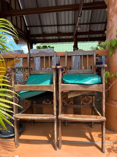 Two weathered wooden chairs, each with a green cushion, are positioned under a covered patio structure in Tambon Ko Panyi, Thailand. The chairs are a central focus, positioned side-by-side. The left chair's cushion is slightly askew. Behind the chairs, decorative wooden paneling and a white, patterned fabric are visible. Above, a corrugated metal roof supported by wooden beams is apparent, with a light fixture hanging from one beam. A fan is mounted on a wooden support to the left. To the left of the chairs, a lush green plant with long, slender leaves in a dark pot stands out. The floor of the patio is an orange, textured surface. A thick, reddish-brown pillar stands to the right, with a green vine growing on it. The background shows glimpses of blue sky and more greenery. The overall scene suggests an outdoor seating area, possibly part of a restaurant or guesthouse, given the rustic and tropical aesthetic.