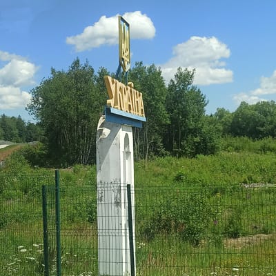 The image shows a signpost for "UKRAINE" in large yellow letters with a blue background, topped by the Ukrainian trident emblem. The signpost is mounted on a white concrete pillar with a carved arch. It is situated in a rural setting with lush green trees and vegetation in the background. A green metal fence runs across the foreground. The sky is blue with scattered white clouds. The location of Vicovu de Sus, Romania, is not directly indicated by any visible elements in the image.