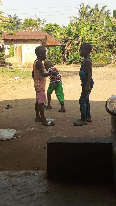 Three children stand in a dirt yard. One child wears a peach-colored tank top and pink shorts, another a striped shirt and green pants, and the third child a blue tank top and jeans. They are near a small house with a rusty roof, surrounded by trees and vegetation. A pair of sandals lies on the ground nearby. The foreground is shadowed, partially obscuring the scene. A bucket sits to the right.