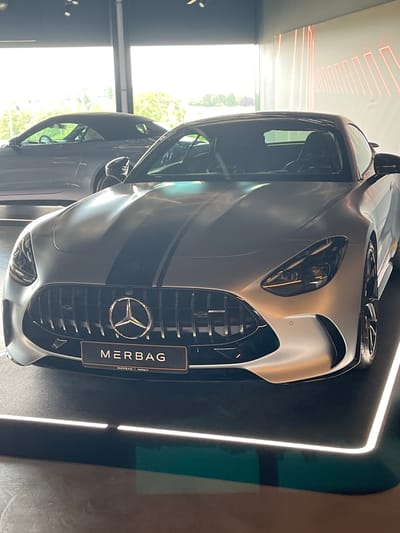 A silver Mercedes-AMG GT Coupe with dual black racing stripes is parked indoors, likely at a dealership or showroom in Reckange-sur-Mess, Luxembourg. The car is positioned prominently in the foreground, facing forward with its grille and headlights clearly visible. A "MERBAG" plaque is affixed below the grille. In the background, another car, possibly a convertible, is partially visible, along with a bright, outdoor-like environment seen through large windows. The interior features a modern aesthetic with red linear lighting accents on the wall and illuminated displays on the floor. The overall impression is a high-end automotive display.