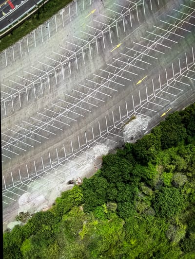 An aerial view of an empty, outdoor parking lot adjacent to a dense, green forest and a road in Vientiane Capital, Laos. The parking lot is paved and marked with white lines indicating parking spaces. Yellow directional arrows are painted on the asphalt. The forest is lush and appears to be a natural landscape bordering the developed area. A portion of a road with yellow and red markings is visible in the upper left corner. There are no people or vehicles present in the parking lot. The scene depicts a functional, yet unused, urban infrastructure element situated next to a vibrant natural environment.